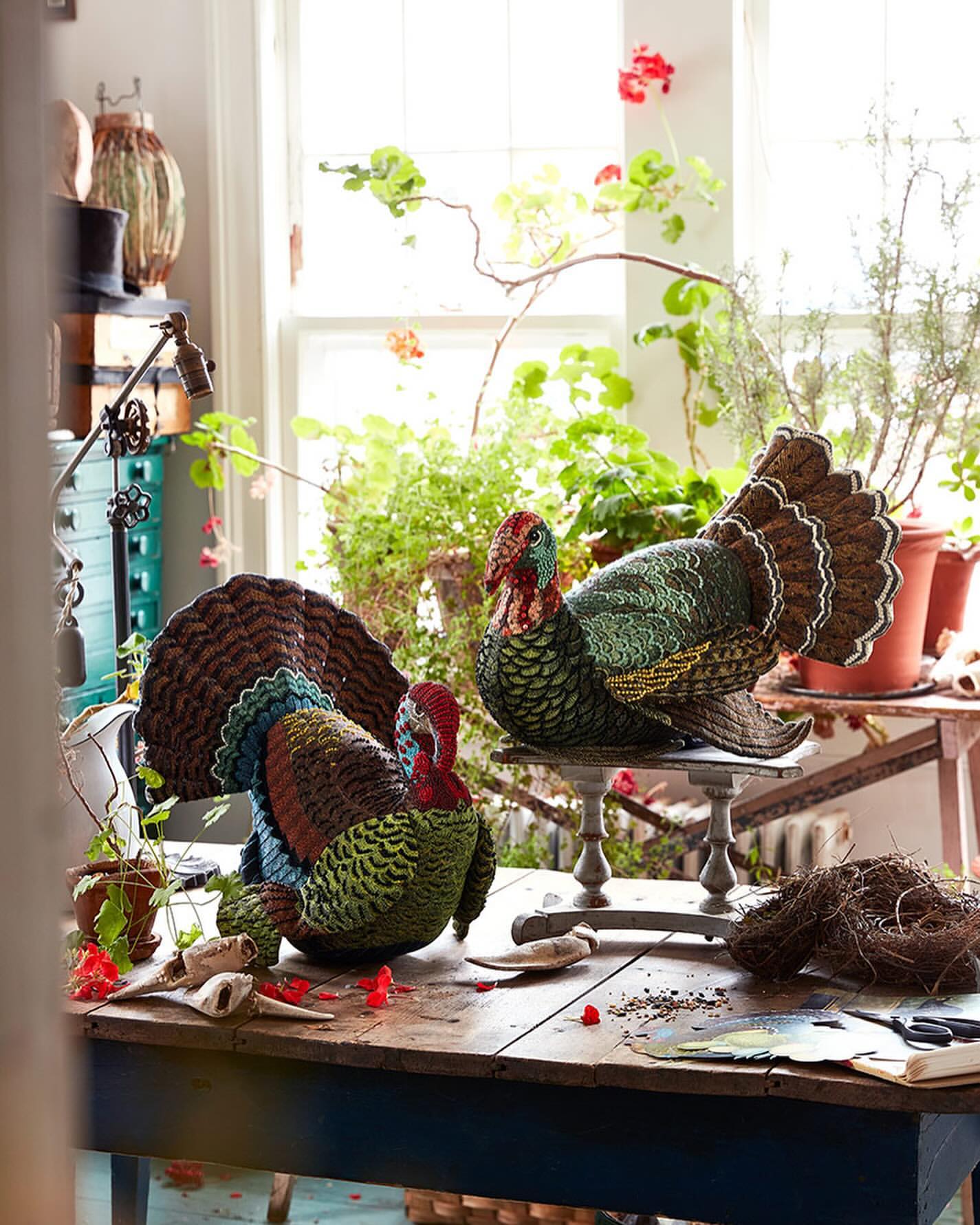 Two decorative turkey sculptures with realistic plumage sit on a rustic wooden table in a naturally lit room filled with plants.