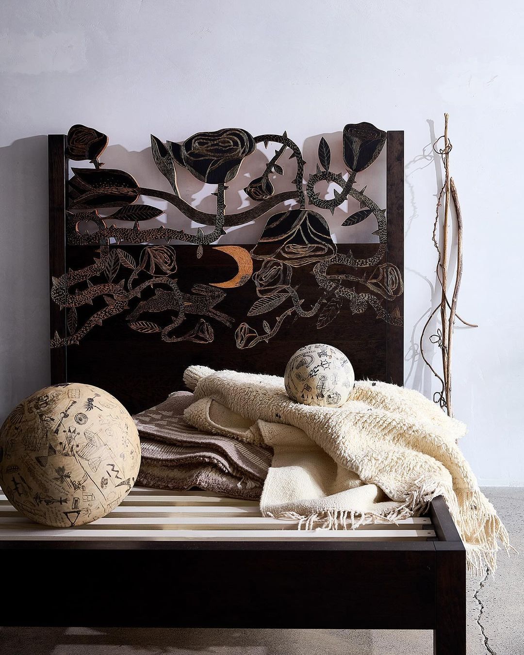 Dark wood bed frame with rose-themed headboard, soft blankets, and spherical patterned cushions in a neutral, shadowed interior.
