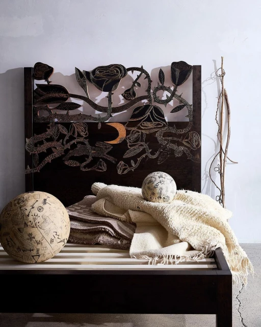Dark wood bed frame with rose-themed headboard, soft blankets, and spherical patterned cushions in a neutral, shadowed interior.