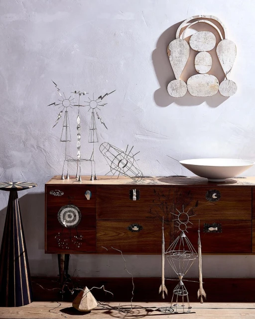 Contemporary art composition featuring geometric wire sculptures, a rustic wooden dresser, white bowl, and a decorative wooden wall hanging.
