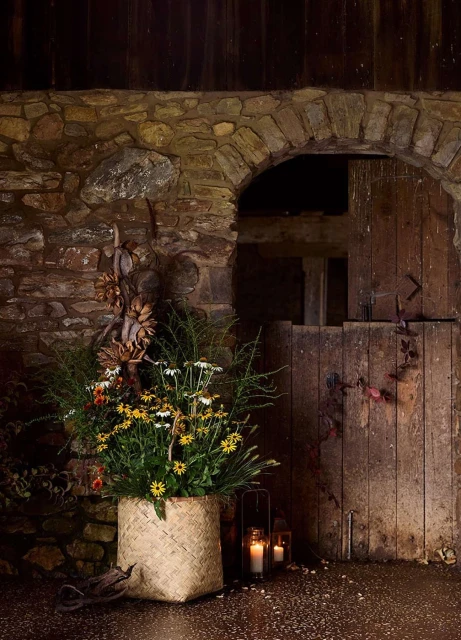 Floral arrangement with daisies and greenery in a woven basket next to lit candle lanterns, set against a stone wall and wood doorway.