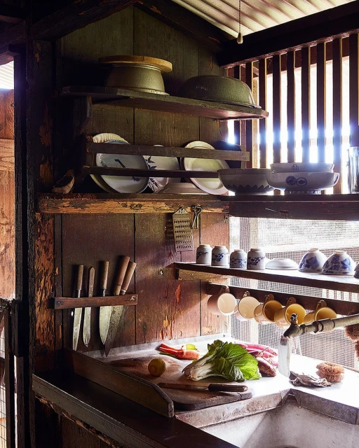 Rustic kitchen interior with wooden shelves holding plates, bowls, cookware, and fresh vegetables on the counter near a sink.