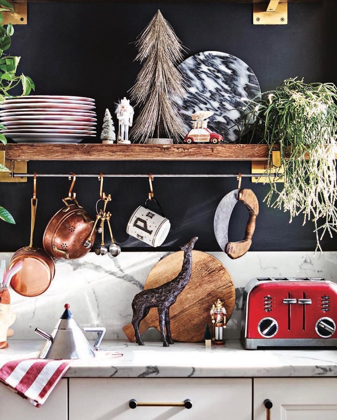 Kitchen still life featuring copper pots, red toaster, cutting boards, holiday decorations, and various kitchenware against dark wall.