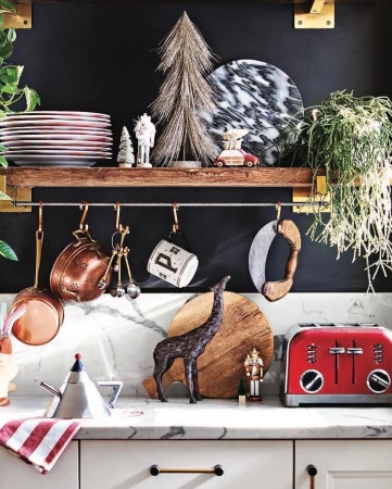 Kitchen still life featuring copper pots, red toaster, cutting boards, holiday decorations, and various kitchenware against dark wall.
