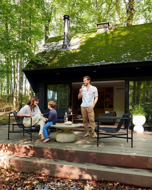 Family relaxes on a wood deck outside a modern cabin surrounded by lush greenery; father drinks wine. Cabin has green mossy roof.