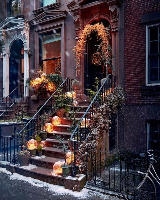 A brownstone exterior is decorated with plants and lights for the holidays, with a bicycle parked along the sidewalk.