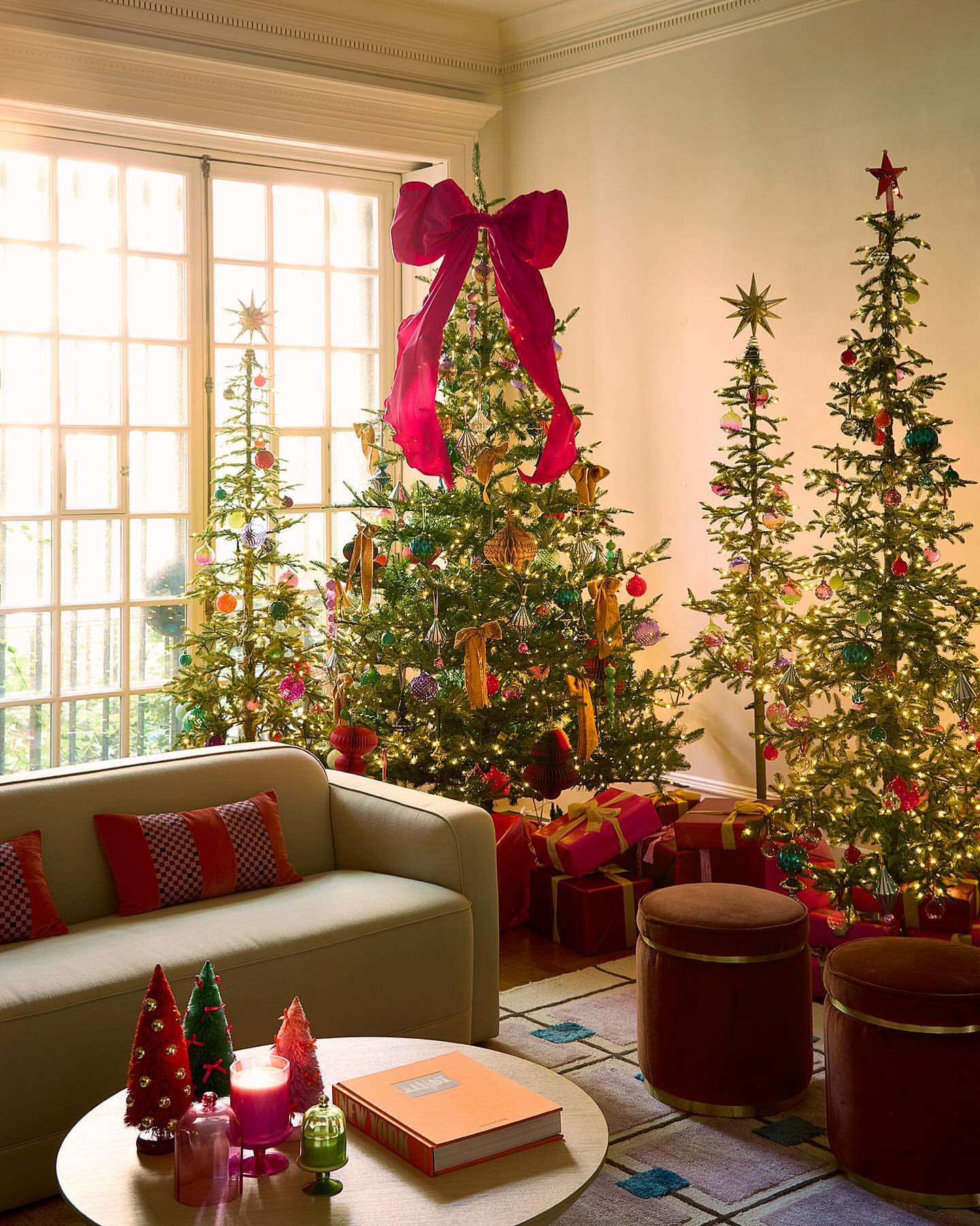 Living room with four decorated Christmas trees, wrapped gifts, a sofa, stools, and a round table with decorative objects.