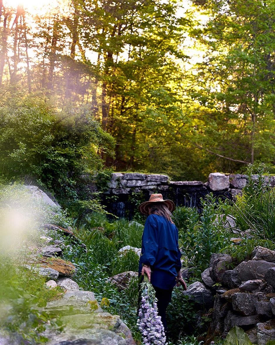 Woman in a hat walks through a verdant garden with a stone wall and a waterfall in the background, bathed in soft, natural light.