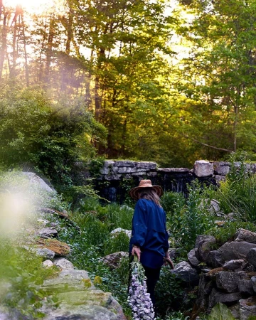 Woman in a hat walks through a verdant garden with a stone wall and a waterfall in the background, bathed in soft, natural light.