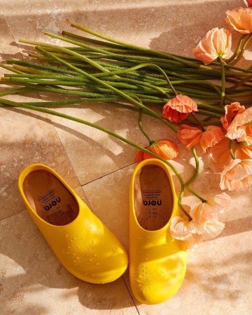 Studio shot of bright yellow slip-on shoes with water droplets alongside peach-colored flowers and green stems on a tan tile surface.
