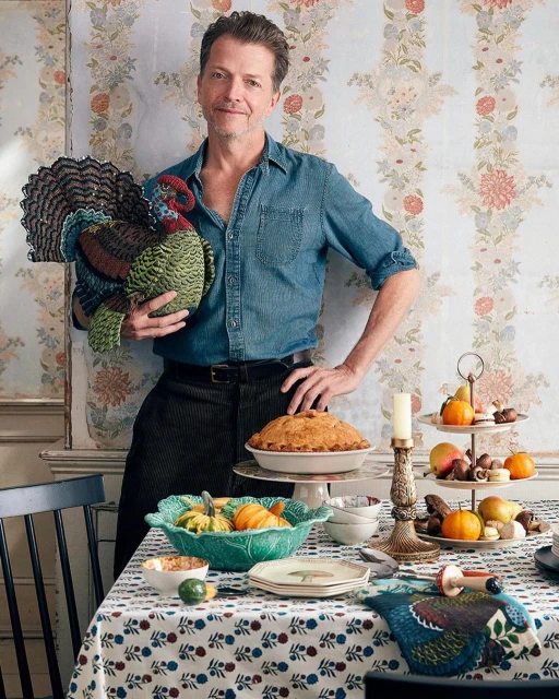 A man in a denim shirt holds a large, decorative turkey next to a table laden with desserts and fall fruits in an indoor setting.