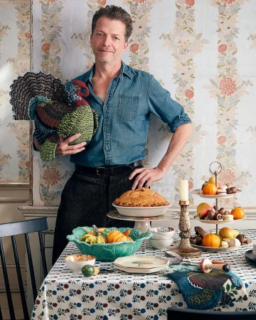 A man in a denim shirt holds a large, decorative turkey next to a table laden with desserts and fall fruits in an indoor setting.
