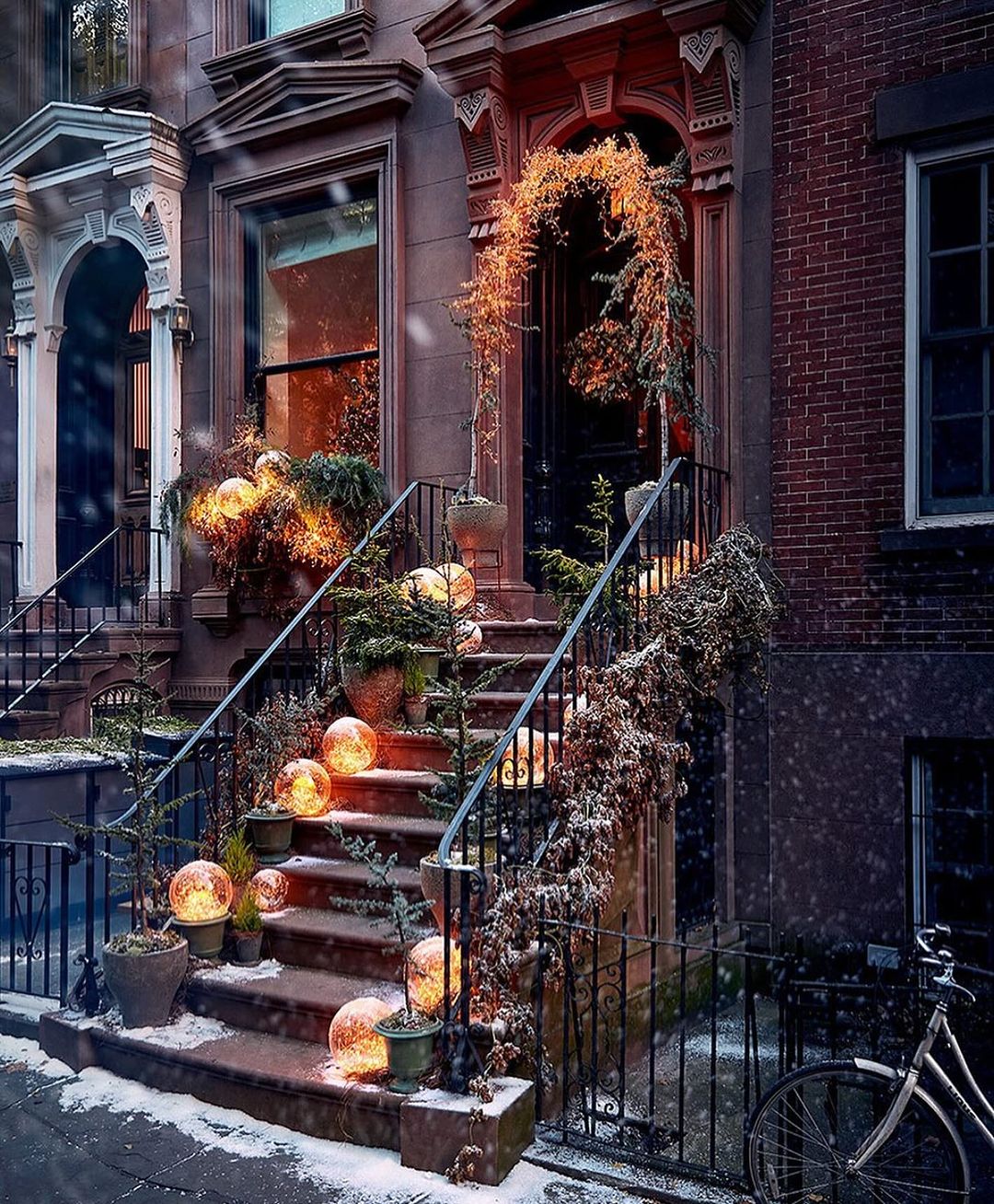 Brownstone entrance adorned with holiday decorations, including illuminated spheres, greenery, and dusted with snow, creating a festive atmosphere.
