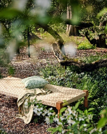 Daybed with a patterned pillow and blanket in a lush green garden setting, with dappled light filtering through the trees.
