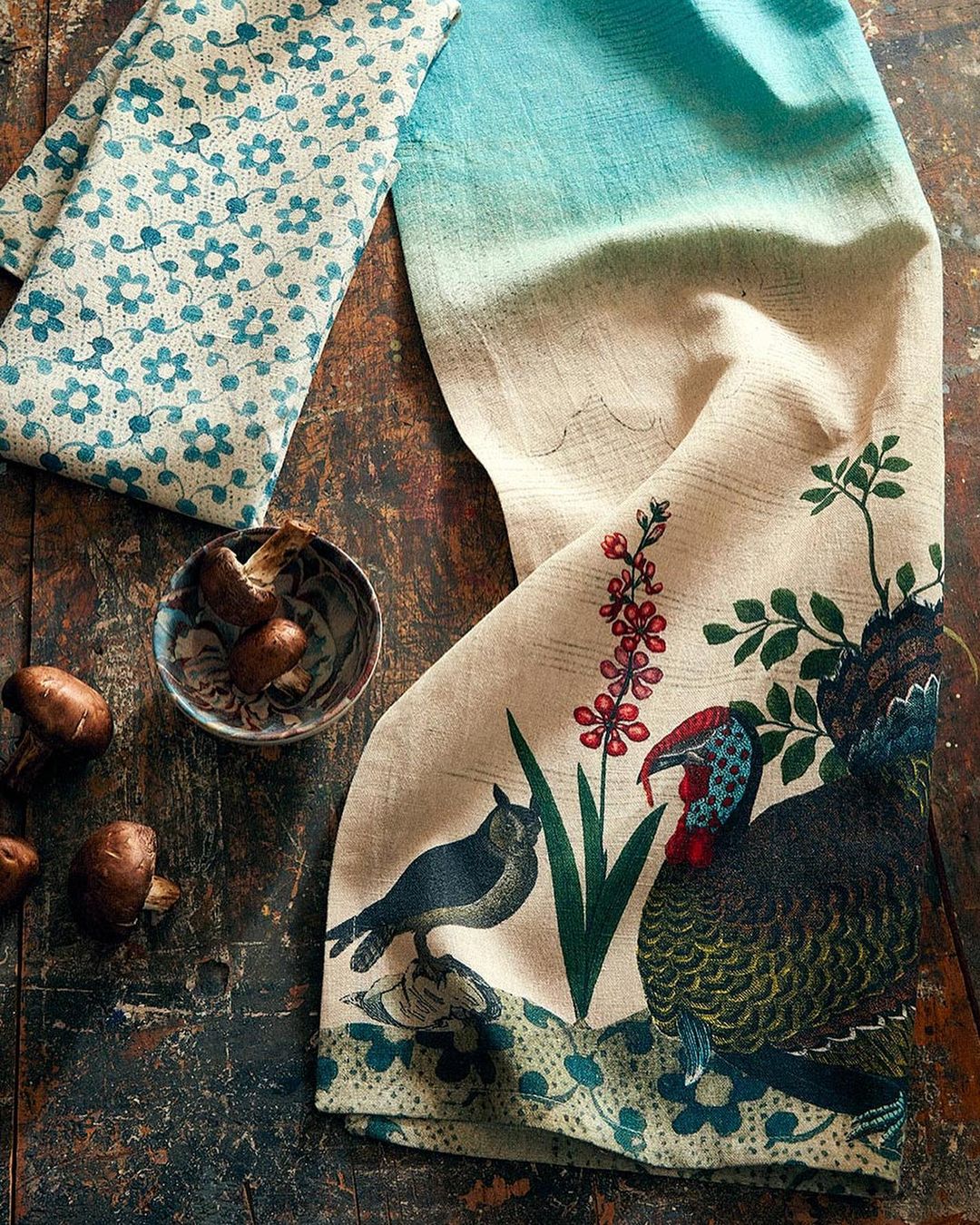 Still life featuring a patterned floral dish towel, a bowl of mushrooms, and printed fabric with animal motifs on a dark wood surface.