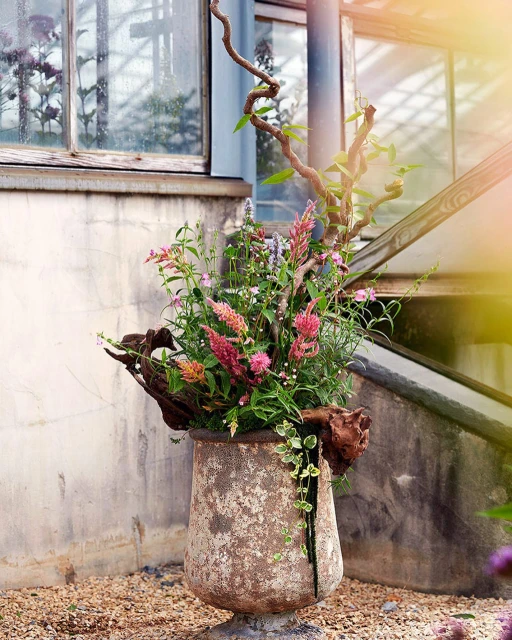 Floral arrangement in aged stone urn features pink and green flowers with winding branches and weathered finish, adding rustic charm.