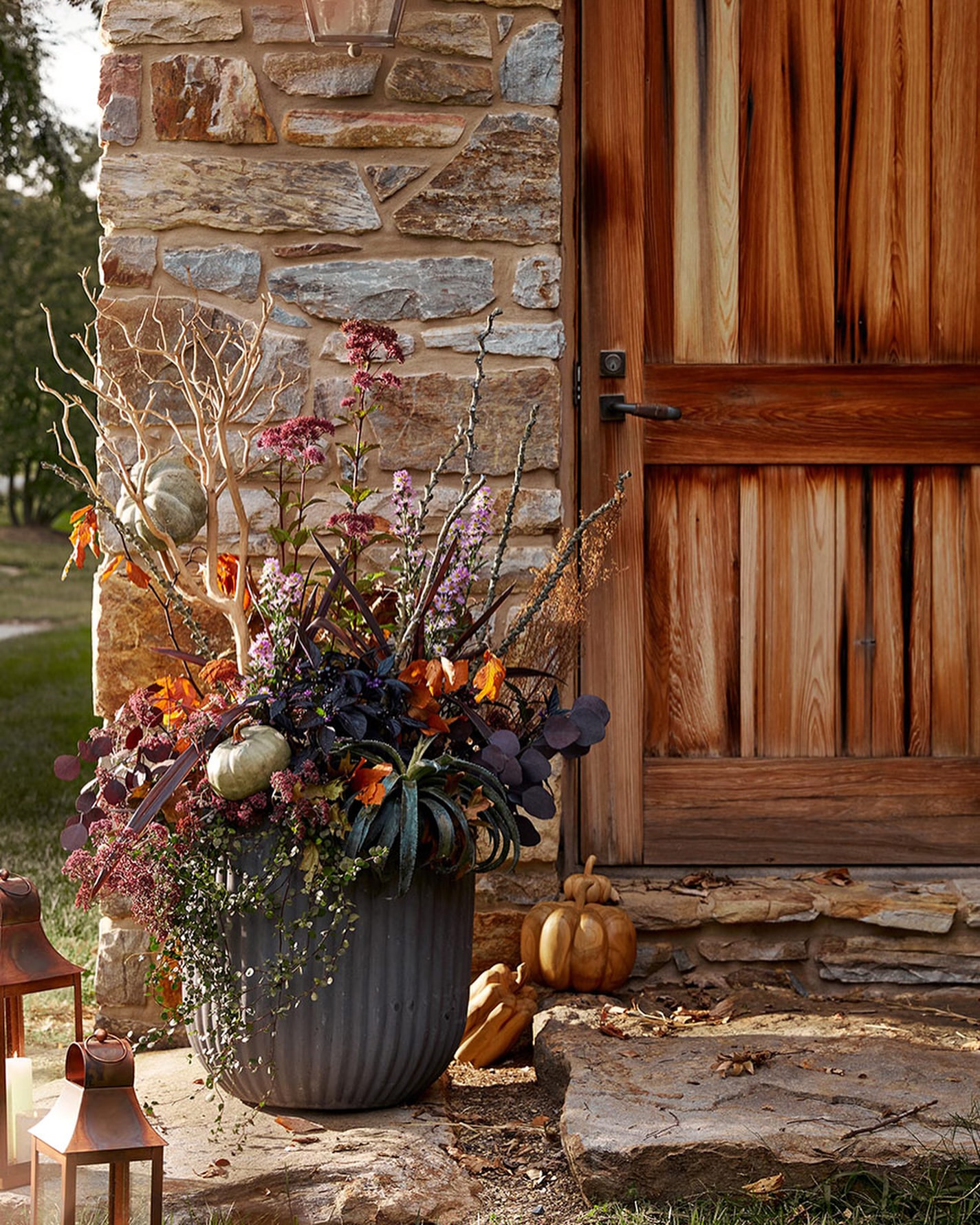 Autumn floral arrangement in a gray planter, featuring pumpkins, leafy plants, branches, and flowers, placed outdoors near a stone wall and door.