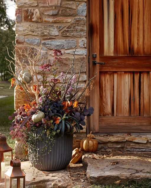 Autumn floral arrangement in a gray planter, featuring pumpkins, leafy plants, branches, and flowers, placed outdoors near a stone wall and door.