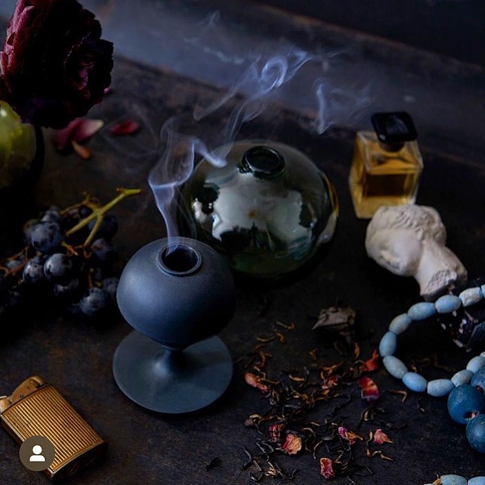 Elegant still life featuring an incense burner emitting smoke, alongside grapes, a perfume bottle, beads, and a bust on a dark surface.