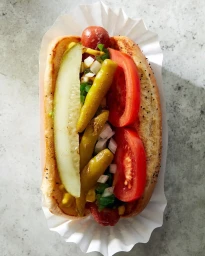 Overhead food photography of a Chicago-style hot dog loaded with tomato wedges, pickle spear, peppers, and onions in a paper holder.