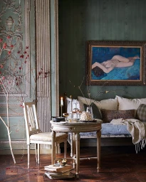 Interior shot of an antique-furnished room with a round wooden table, white chair, and an oil painting hanging on a dark green wall.