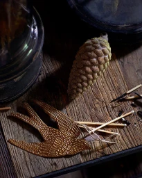 Still life arrangement with a golden swallow ornament, a pinecone, and matches on a rustic wooden table by Dane Tashima.