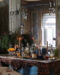 A laden marble-topped table with wine bottles, candles, fruit, and foliage beneath an ornate mirror by Dane Tashima