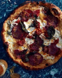 Wood-fired pizza with dark cured meat, melted white cheese, and dark basil leaves on a blue patterned surface.