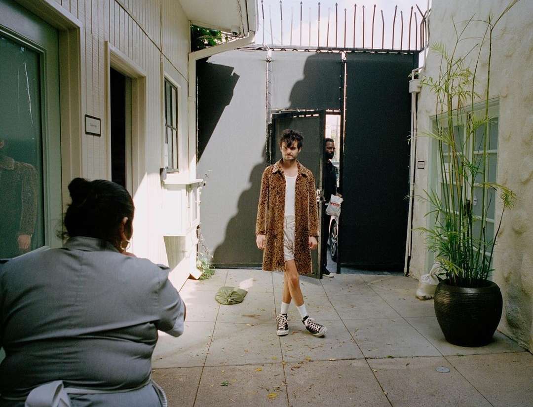 An outdoor fashion shoot features a model in a cheetah print coat, white shorts, and sneakers, posed in front of black gates.