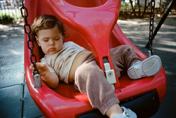 A toddler sleeping soundly inside a bright red plastic baby swing at an outdoor playground in New York.