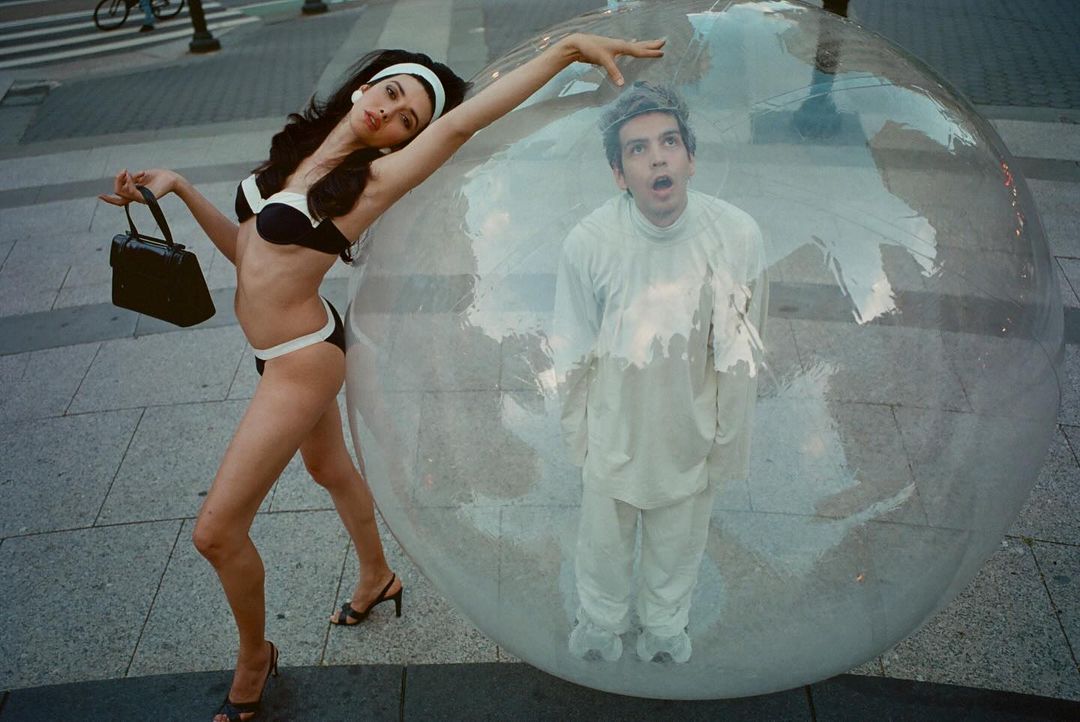 Woman in a black and white bikini posing next to a large plastic bubble containing a man in white clothing on a paved street.