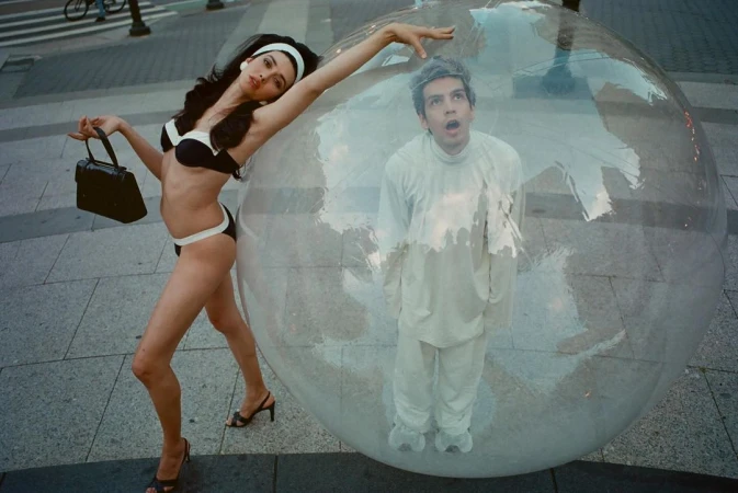 Woman in a black and white bikini posing next to a large plastic bubble containing a man in white clothing on a paved street.