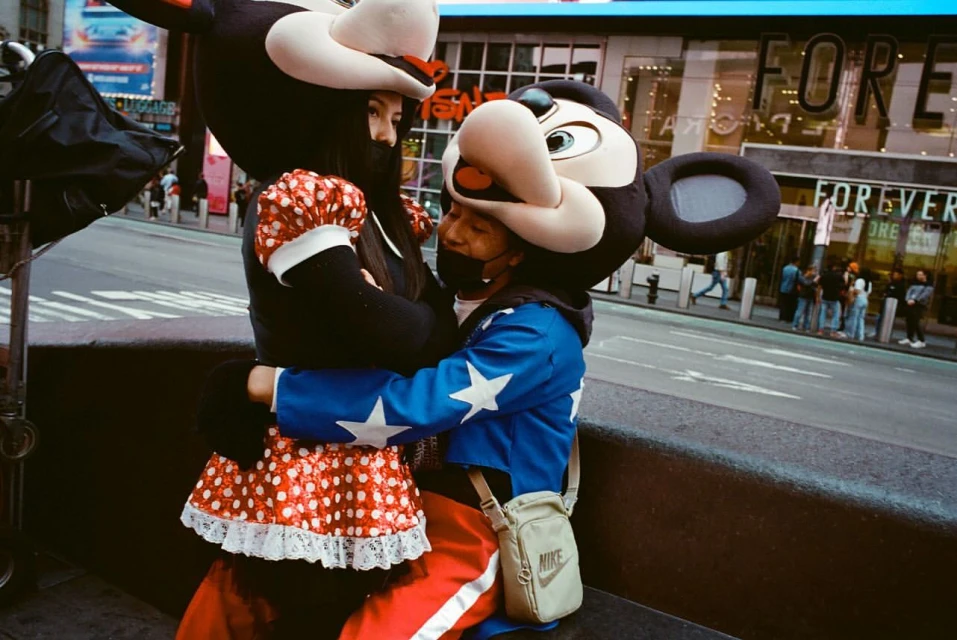 Two people in mouse mascot costumes embrace on a city street with a Forever 21 store in the background and crosswalk in view.