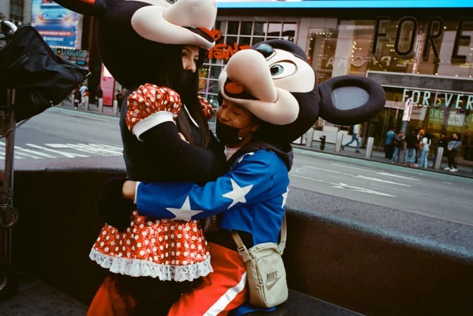 Two people in mouse mascot costumes embrace on a city street with a Forever 21 store in the background and crosswalk in view.