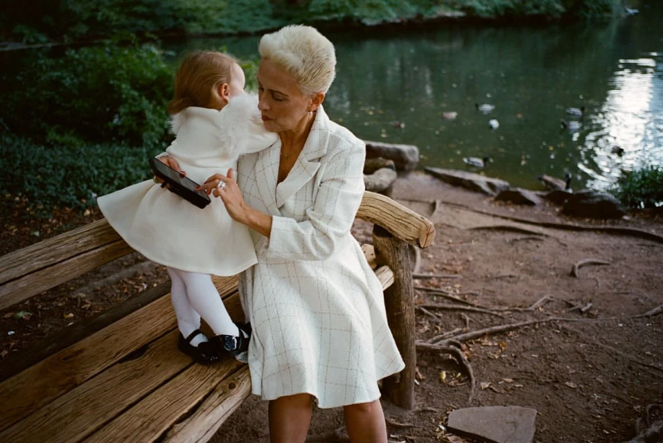 Adult woman and child on a wooden bench in a park with a pond. The woman wears a cream-colored coat and the child wears a white dress.