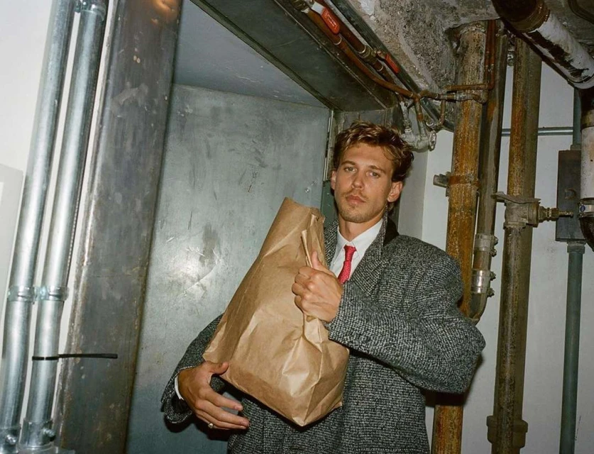 A man in a coat and tie standing in a basement holding a paper bag, next to metal pipes and an open doorway, looking at the camera.