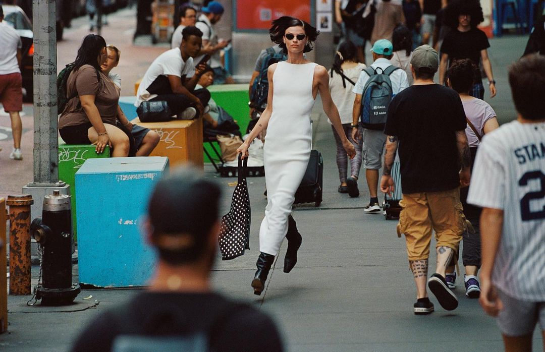 Fashionable adult woman in a white dress walking on a city street surrounded by pedestrians and urban elements on a daytime.