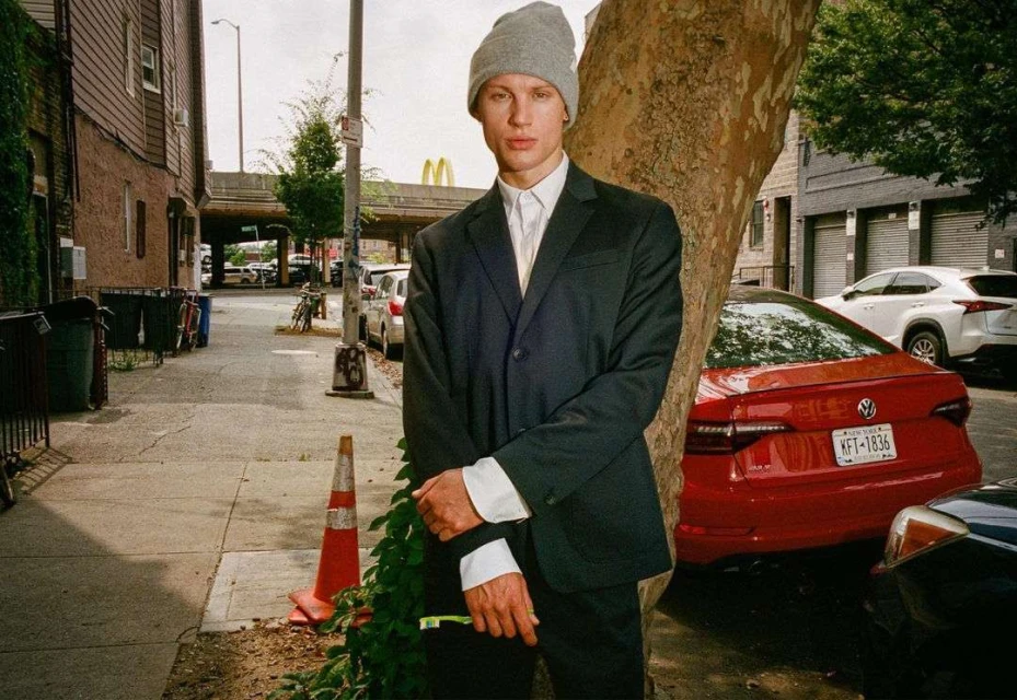 Adult man in a suit and grey beanie standing on a city street next to a tree, with cars and buildings in the background.