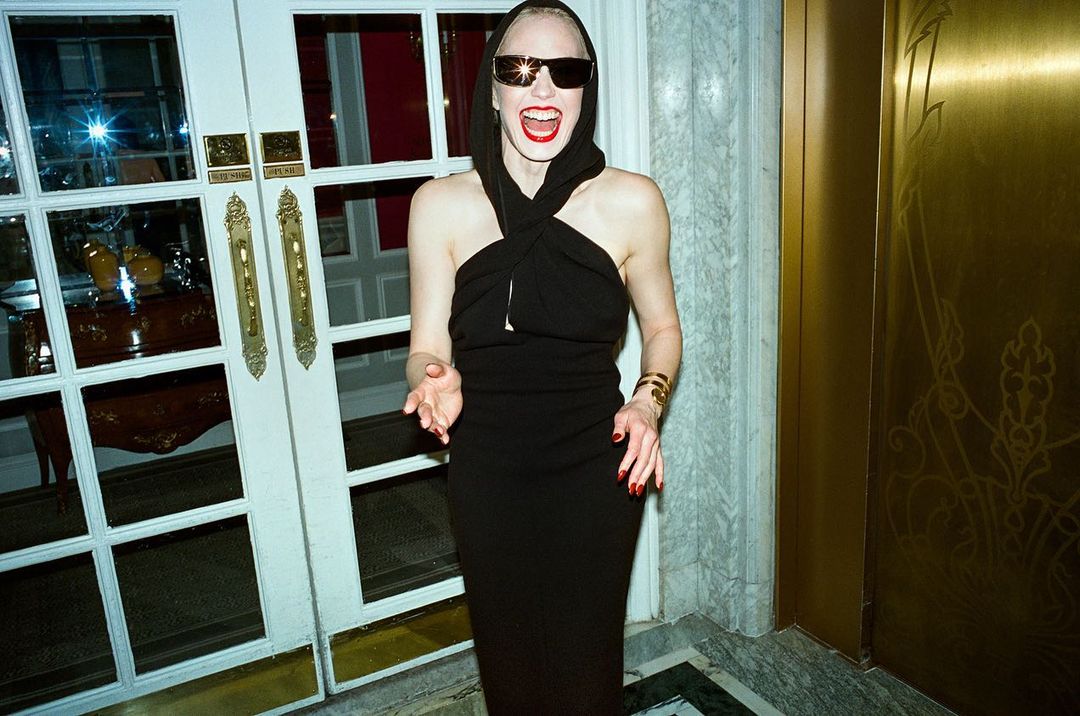 Woman in black dress and sunglasses, smiling with open mouth, stands inside doorway. Gold door and white-frame glass doors visible.