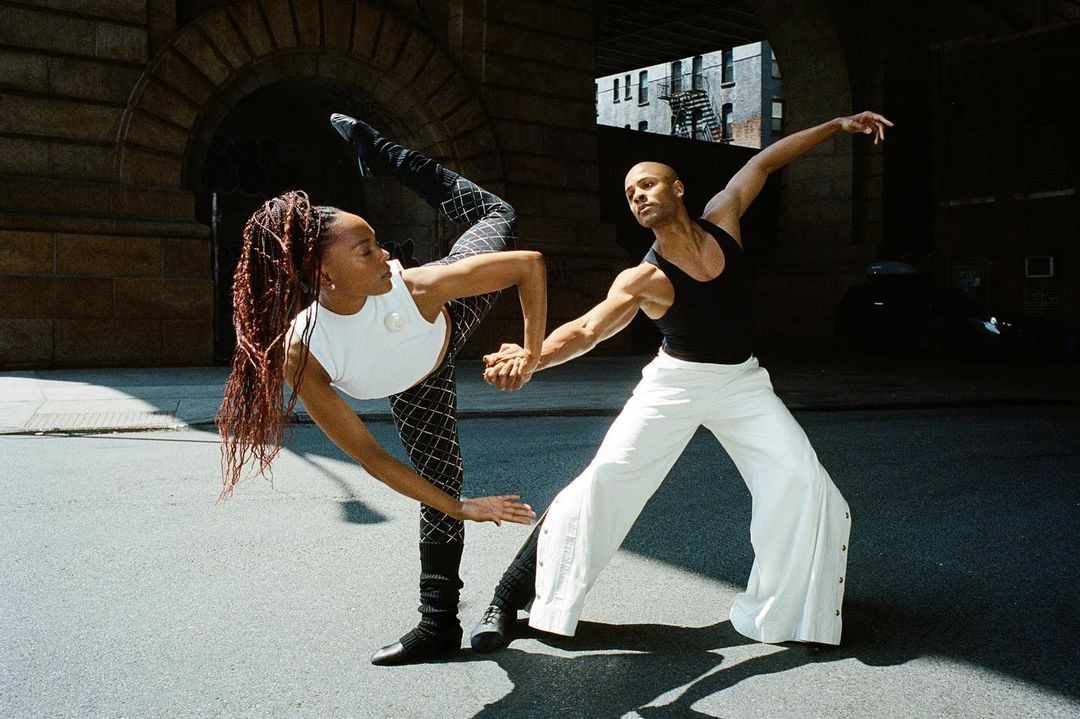 Two dancers, one Black woman with braids and one bald man, perform together on a city street under a brick archway.