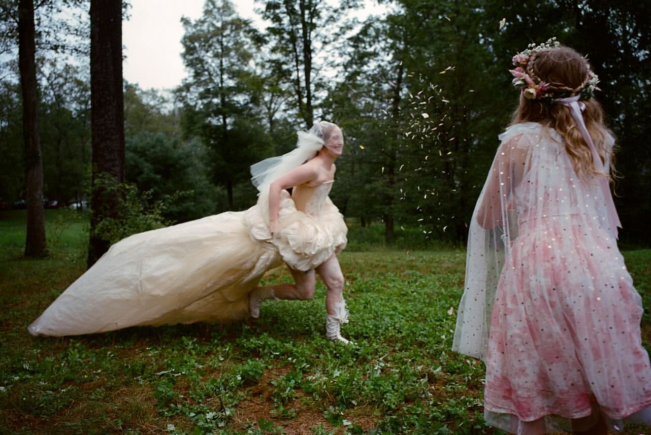 A person in a wedding dress runs through a grassy field as flower petals are thrown toward them by a person with a floral crown.