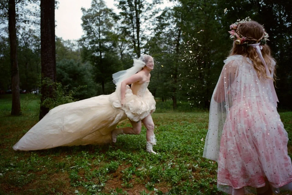 A person in a wedding dress runs through a grassy field as flower petals are thrown toward them by a person with a floral crown.