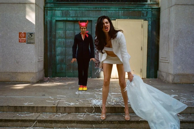 Woman in white suit shouts while holding a discarded veil on steps with a devil-costumed person in background outdoors.