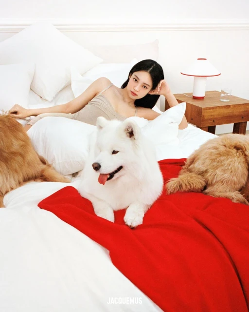 An adult woman poses on a white bed next to a white Samoyed and a reddish-brown dog; a red blanket adds a pop of color.