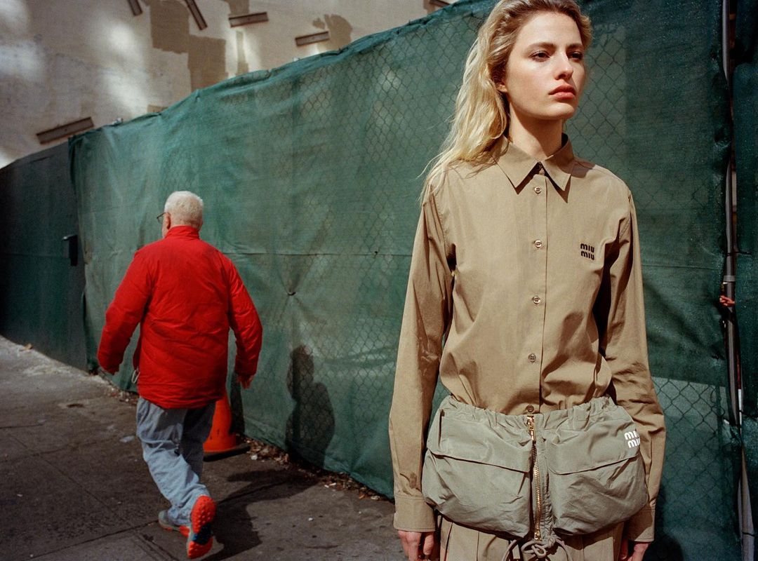 Fashion shot of a fair-skinned woman in a beige shirt and matching skirt, posing on a city street near a chain-link fence.
