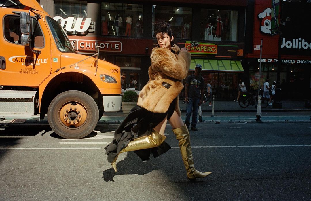 A stylish adult woman in a fur coat and gold thigh-high boots confidently strides across a city street, next to an orange truck.