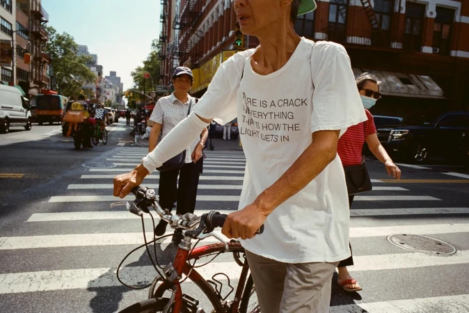 Older woman on bicycle crosses street in urban setting, wearing white t-shirt with quote, accompanied by pedestrians and vehicles.