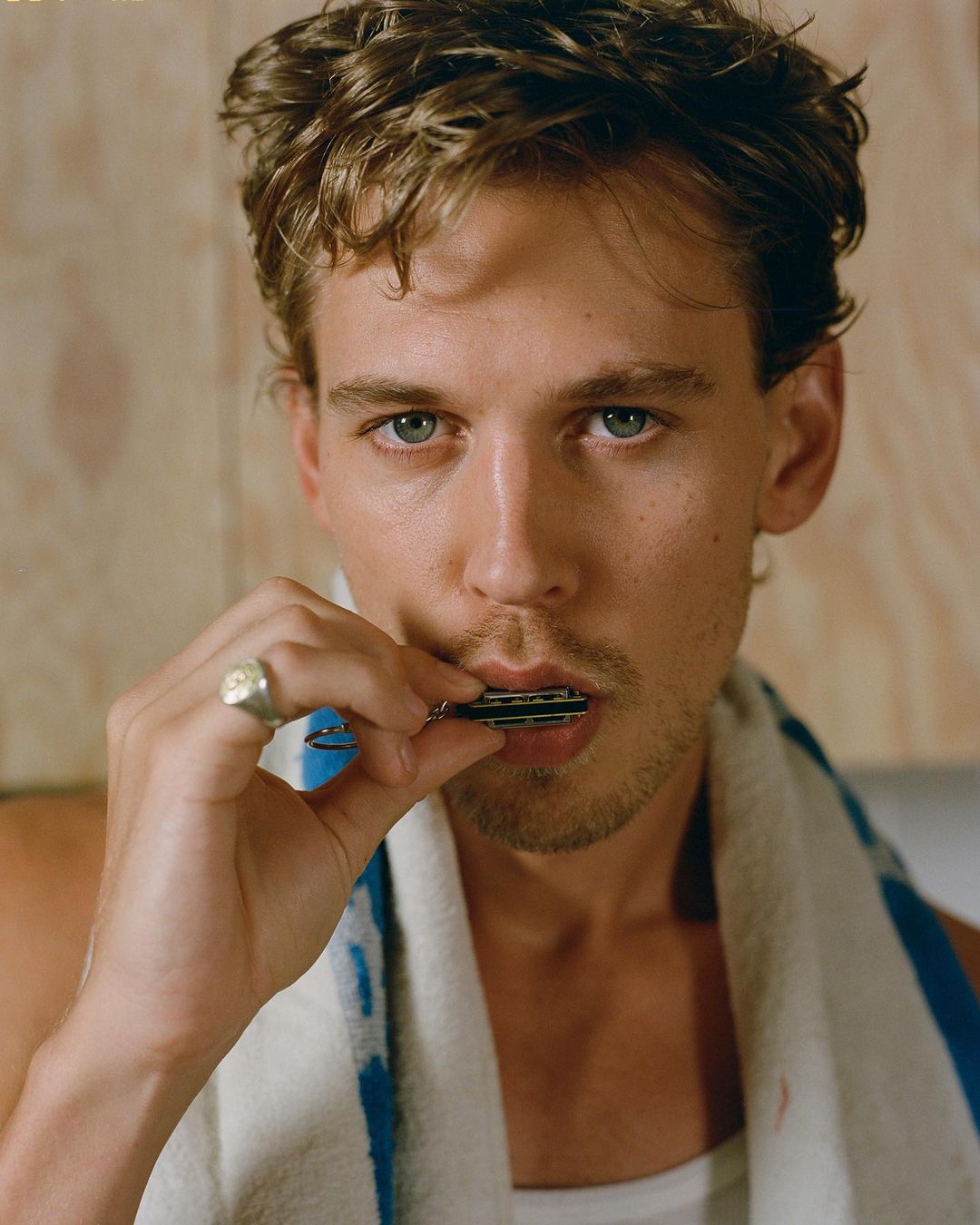 Adult male with wavy hair and a silver ring is standing indoors in front of a light-colored wall, brushing his teeth with a white towel nearby.