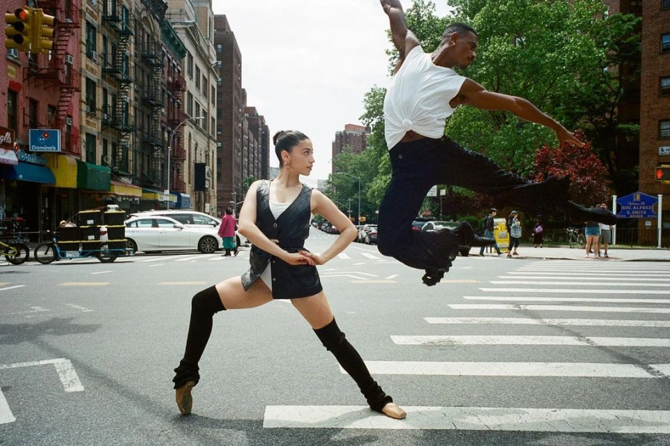 Two dancers, an adult female and an adult male, performing ballet moves on a city street with apartment buildings in the background.