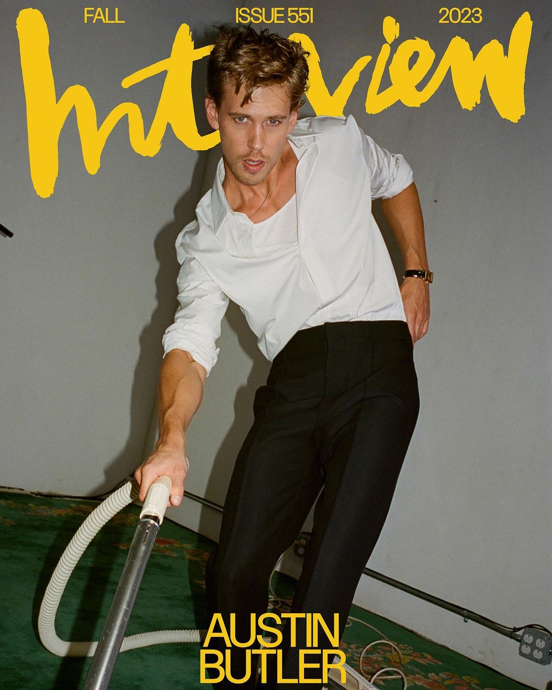 Austin Butler poses with a vacuum cleaner on a magazine cover. The backdrop is a gray wall and a green carpet.